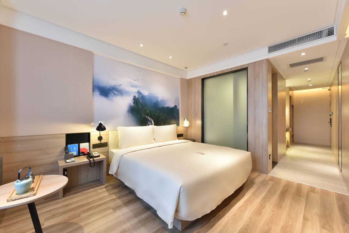 Une chambre d’hôtel moderne dotée d’un grand lit, d’une table de chevet, d’une lampe et offrant une vue sur une montagne grâce à un mur en verre.