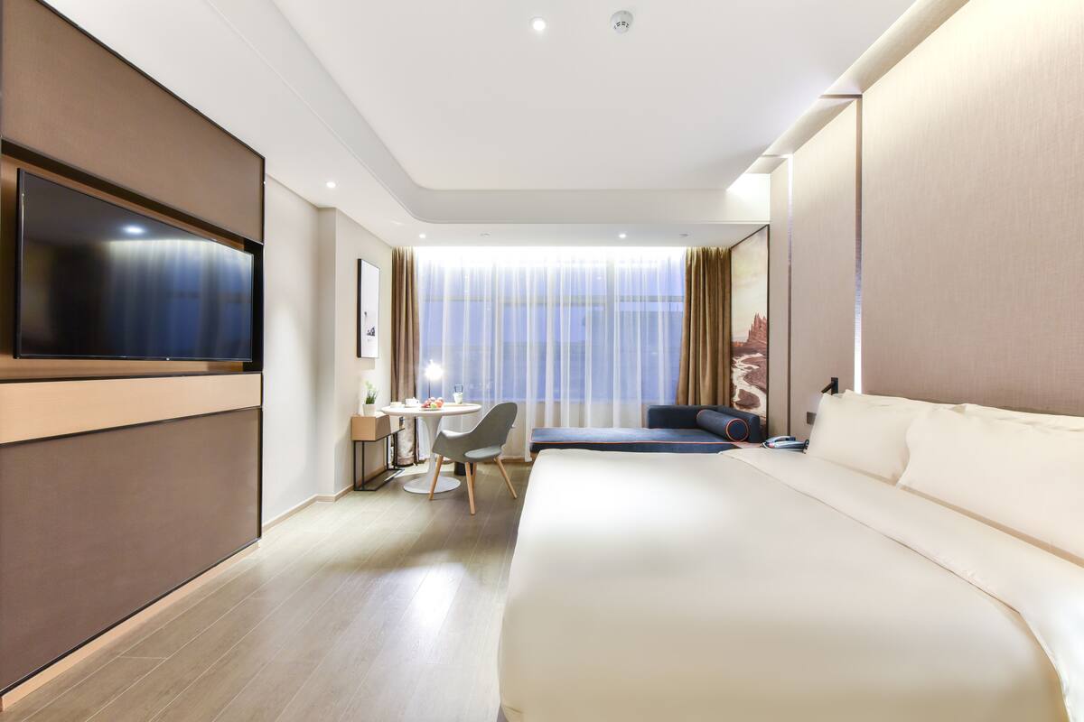 A modern hotel room with a large bed, a desk with a chair, a flat-screen TV, and a large window with sheer curtains.