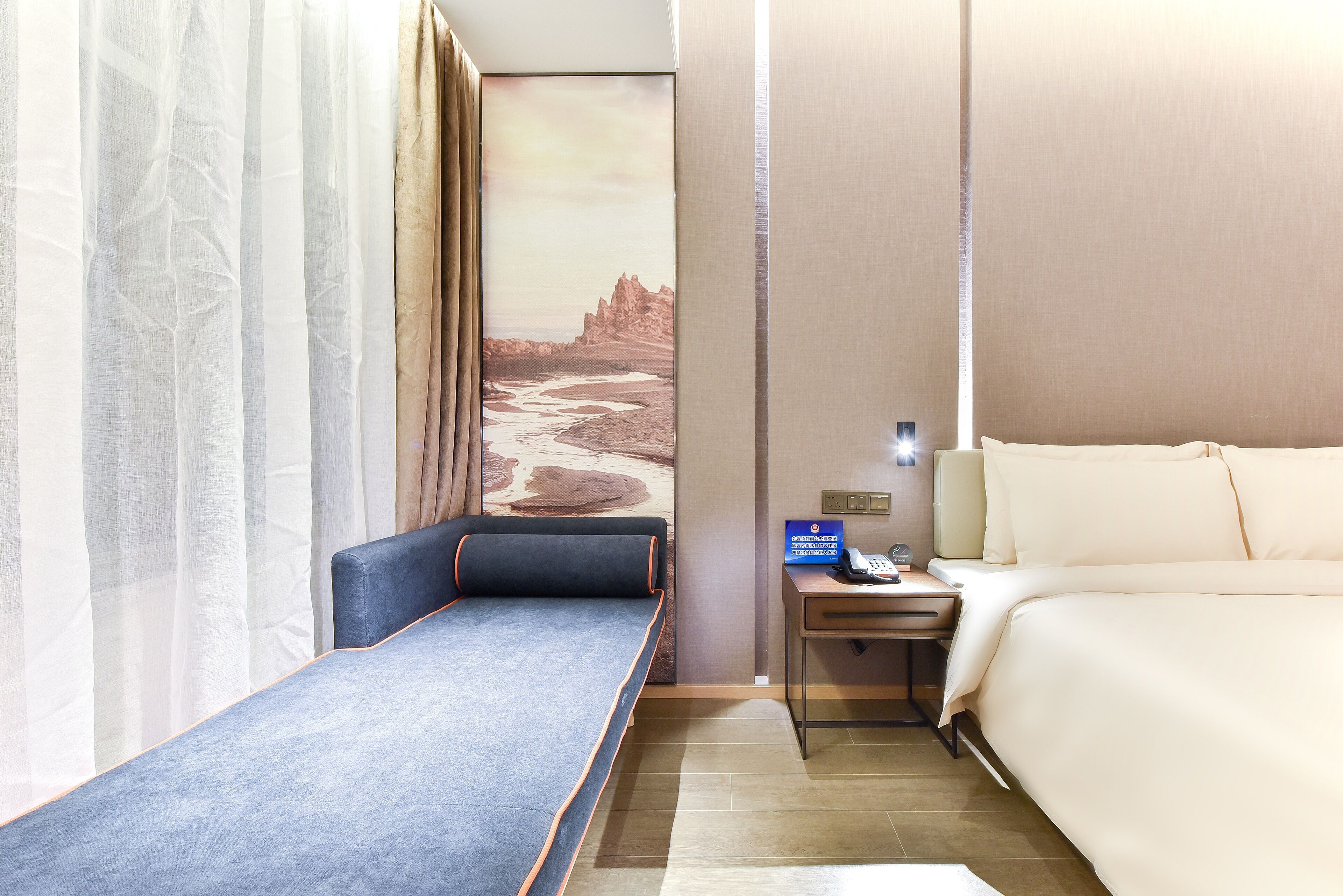 A hotel room with a single bed, a small sofa, a nightstand with a lamp, and a painting of a desert landscape on the wall.