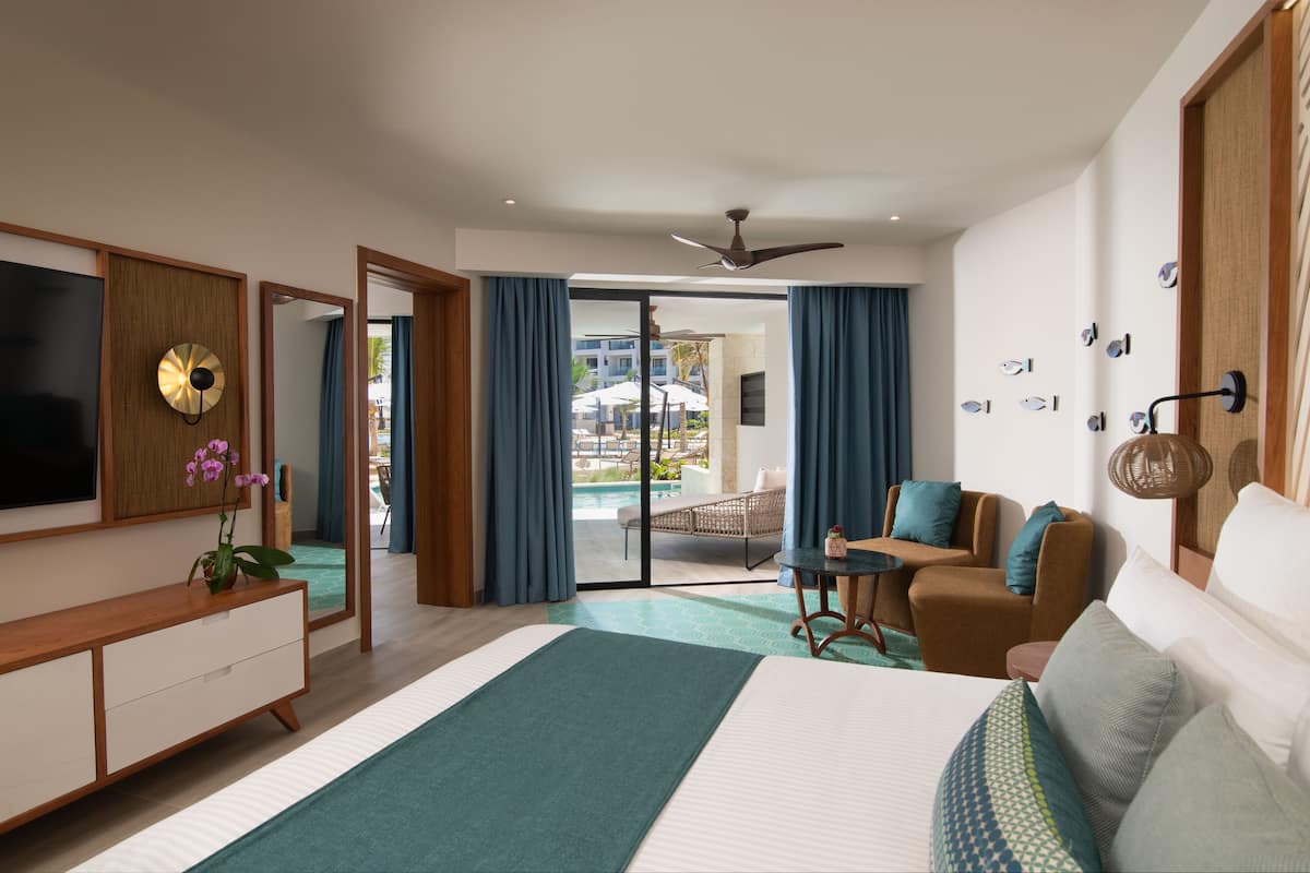 A modern hotel room with a large bed, a sofa, a TV, and a view of the pool and outdoor area.