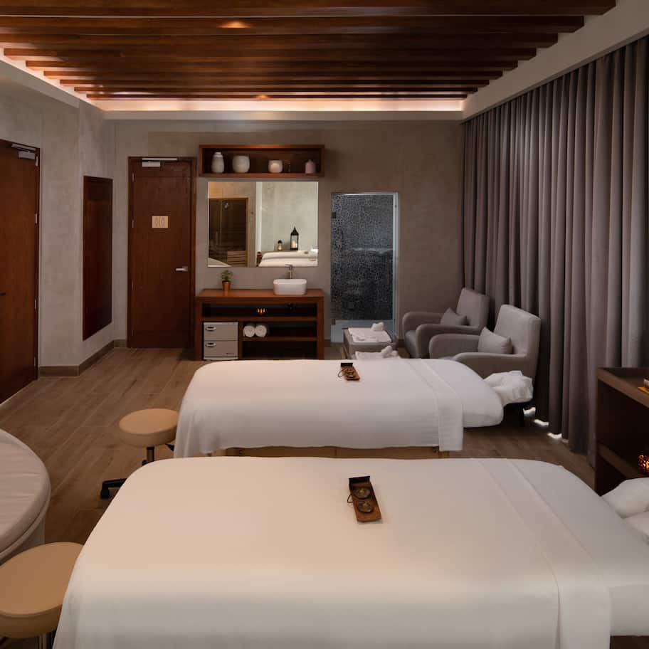A modern hotel room with two beds, a vanity with a mirror, a small seating area, and a large window with curtains.
