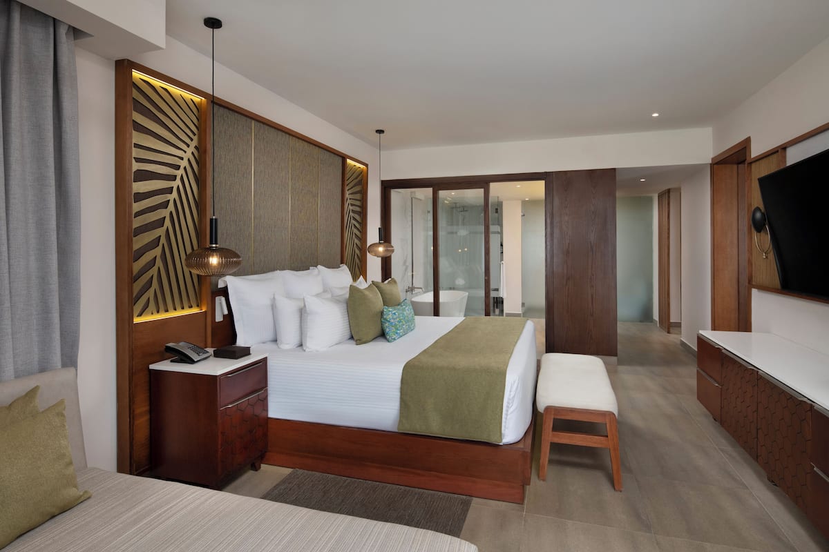 A modern hotel bedroom with a large bed, bedside tables, a bench, a TV, and a sliding glass door leading to a bathroom.
