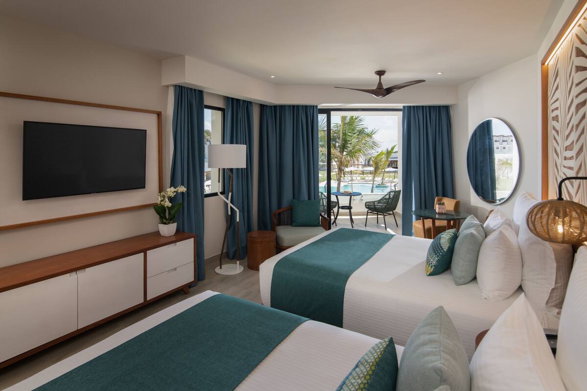 A hotel room with two beds, a TV, a desk, and a view of a pool and palm trees.