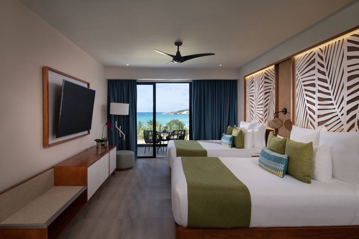 A modern hotel room with two beds, a flat-screen TV, a desk, and a view of the sea.