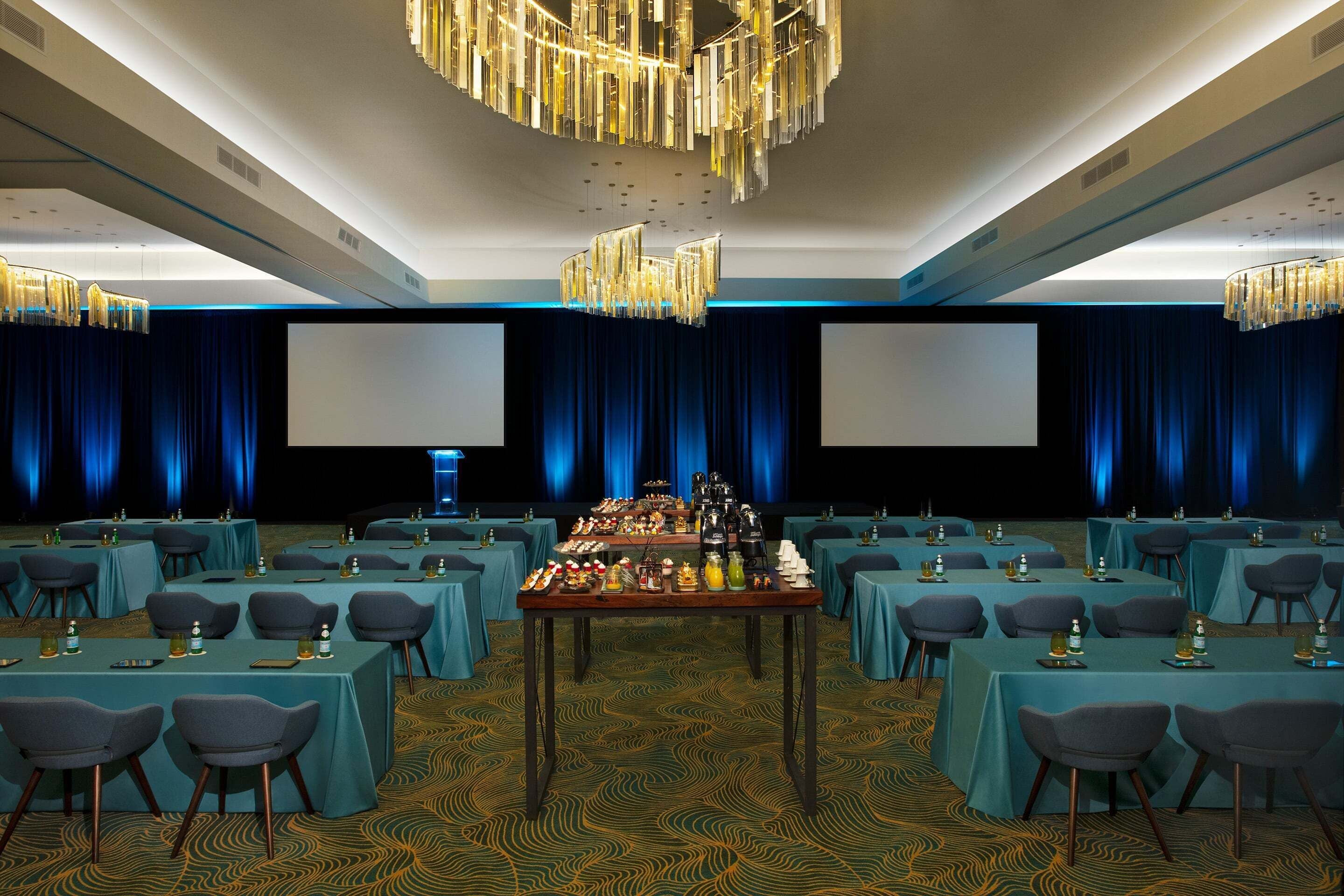 A conference room with tables set for a meeting, a large screen, and a chandelier.