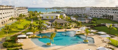 A resort with a central swimming pool, multiple buildings, and a view of the ocean.