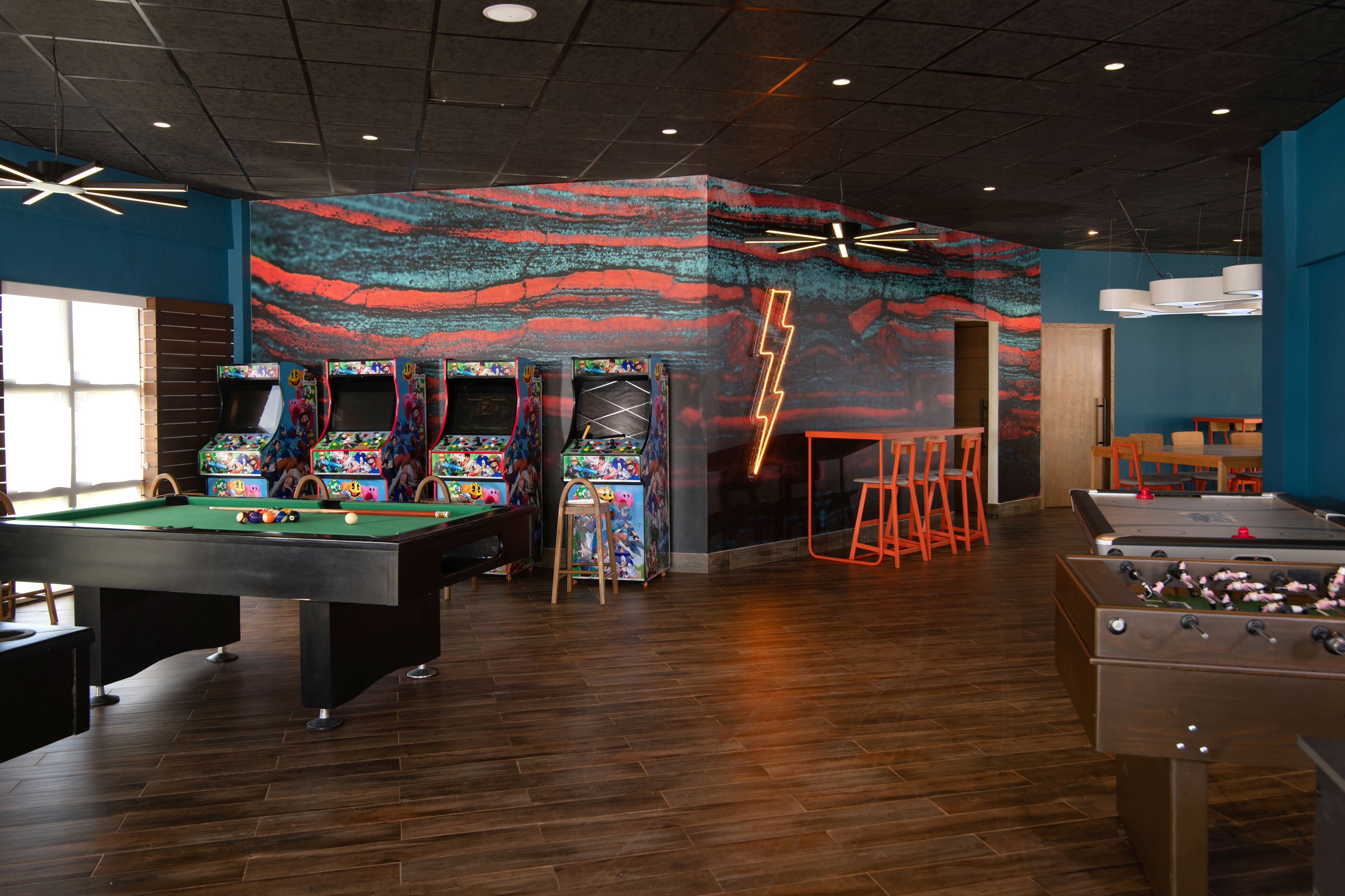 A pool table, foosball table, and arcade games in a room with a wooden floor and blue walls.