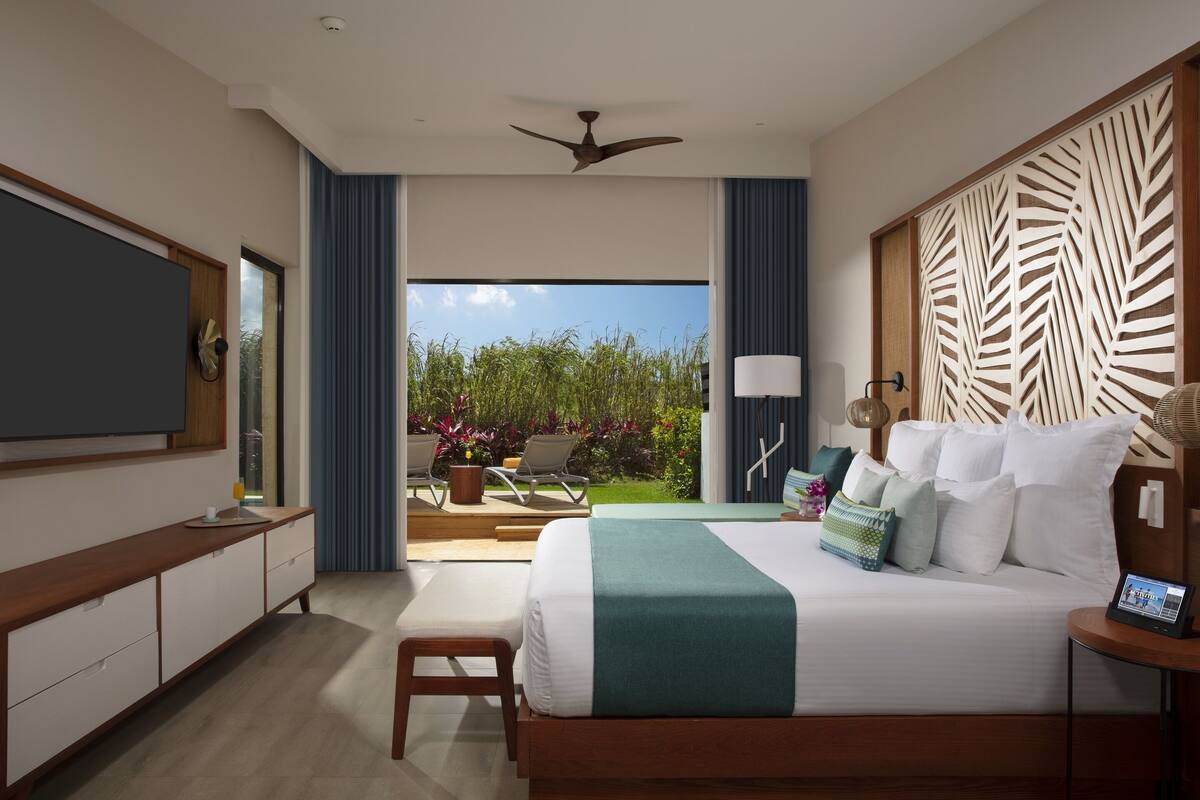 A modern hotel room with a large bed, a TV, a desk, and a balcony with a view of greenery.