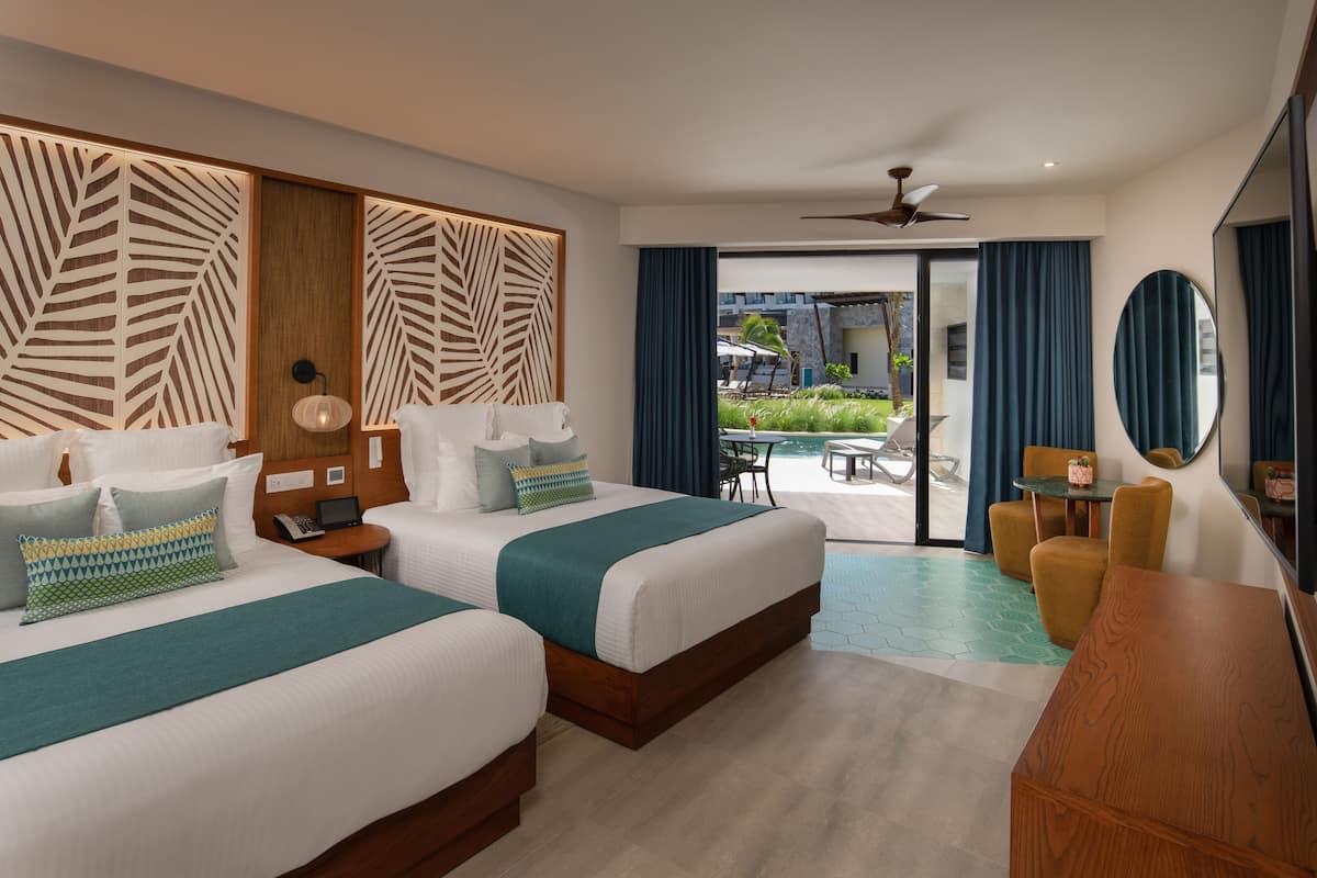 A hotel room with two beds, a desk, a chair, a ceiling fan, and a view of the outdoor area.