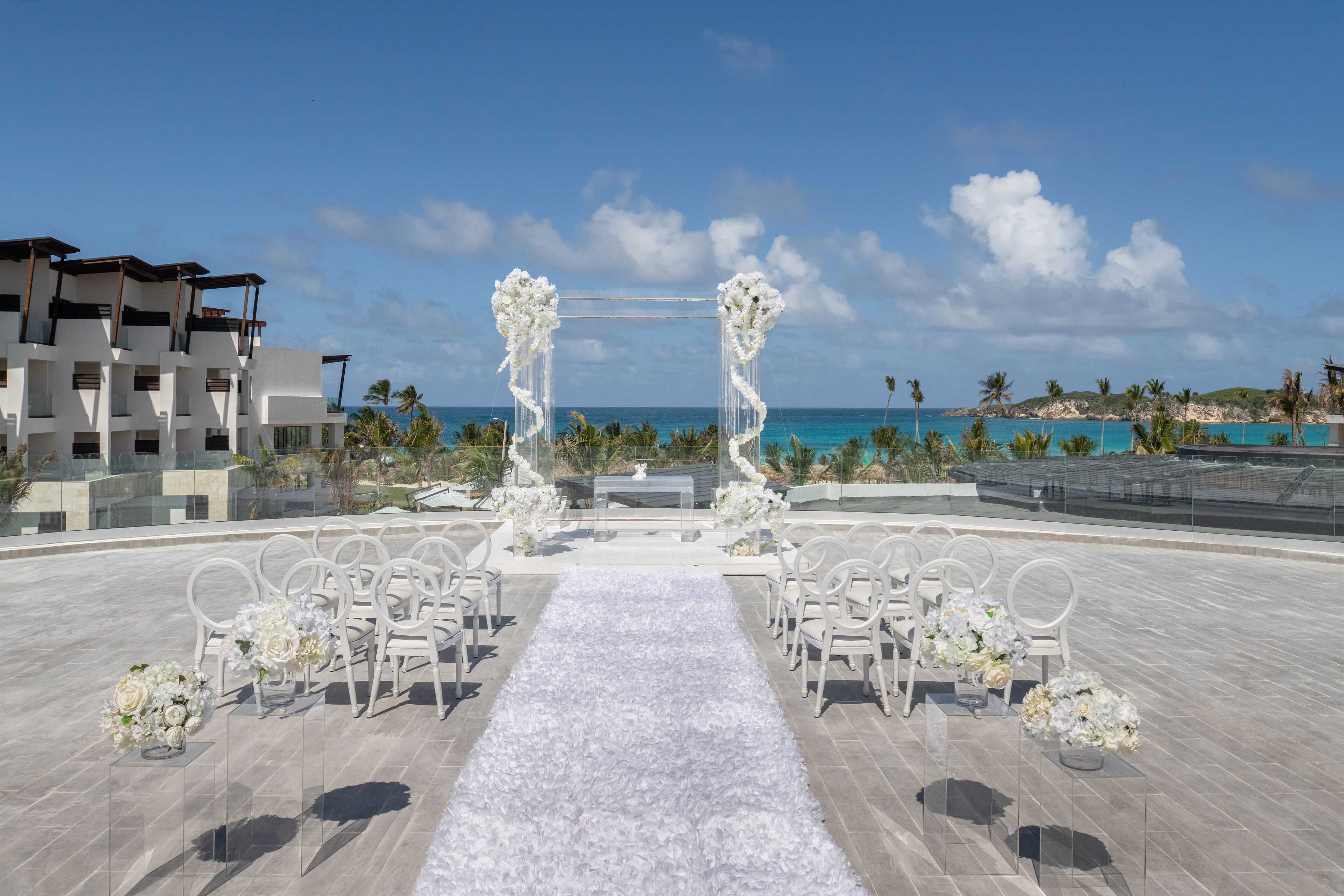 Ceremonia de boda al aire libre con sillas blancas y decoraciones florales, con vista a una playa y al océano.