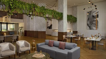A modern hotel lobby with a grey sofa, armchairs, a glass coffee table, and a dining area with tables and chairs.