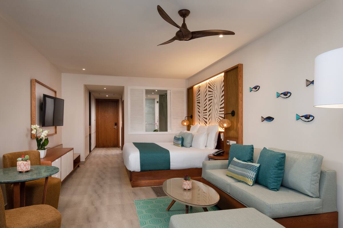 A modern hotel room with a bed, sofa, dining area, and a TV.