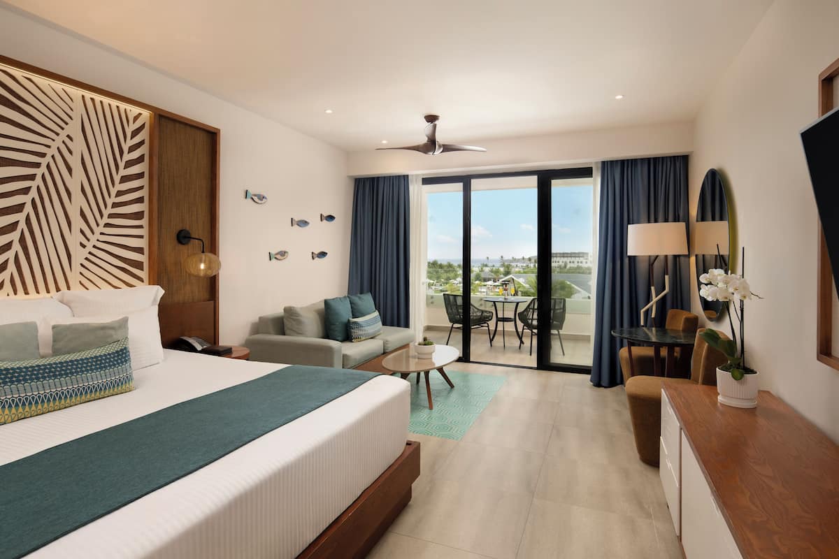 A hotel room with a large bed, a sofa, a dining table, and a balcony with a view.