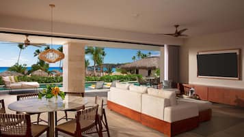 A modern living room with a large sofa, a dining table, and chairs. There is a view of a pool and palm trees outside.