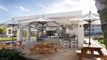 Un patio al aire libre con mesas de picnic de madera y sombrillas blancas, una zona de bar y vistas a un edificio moderno al fondo.
