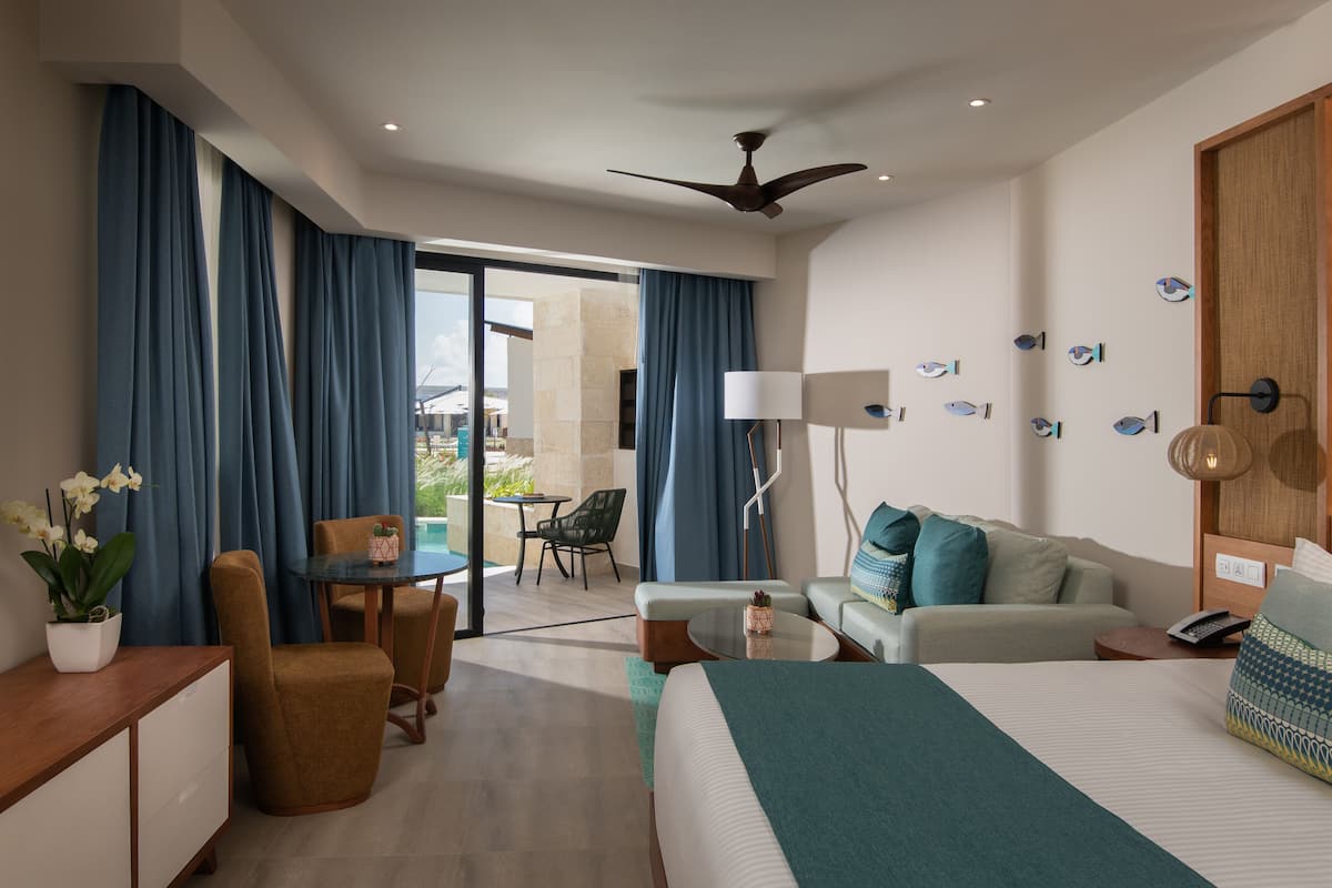 A modern hotel room with a bed, sofa, dining table, and chairs. There is a balcony with a view of the outdoors.