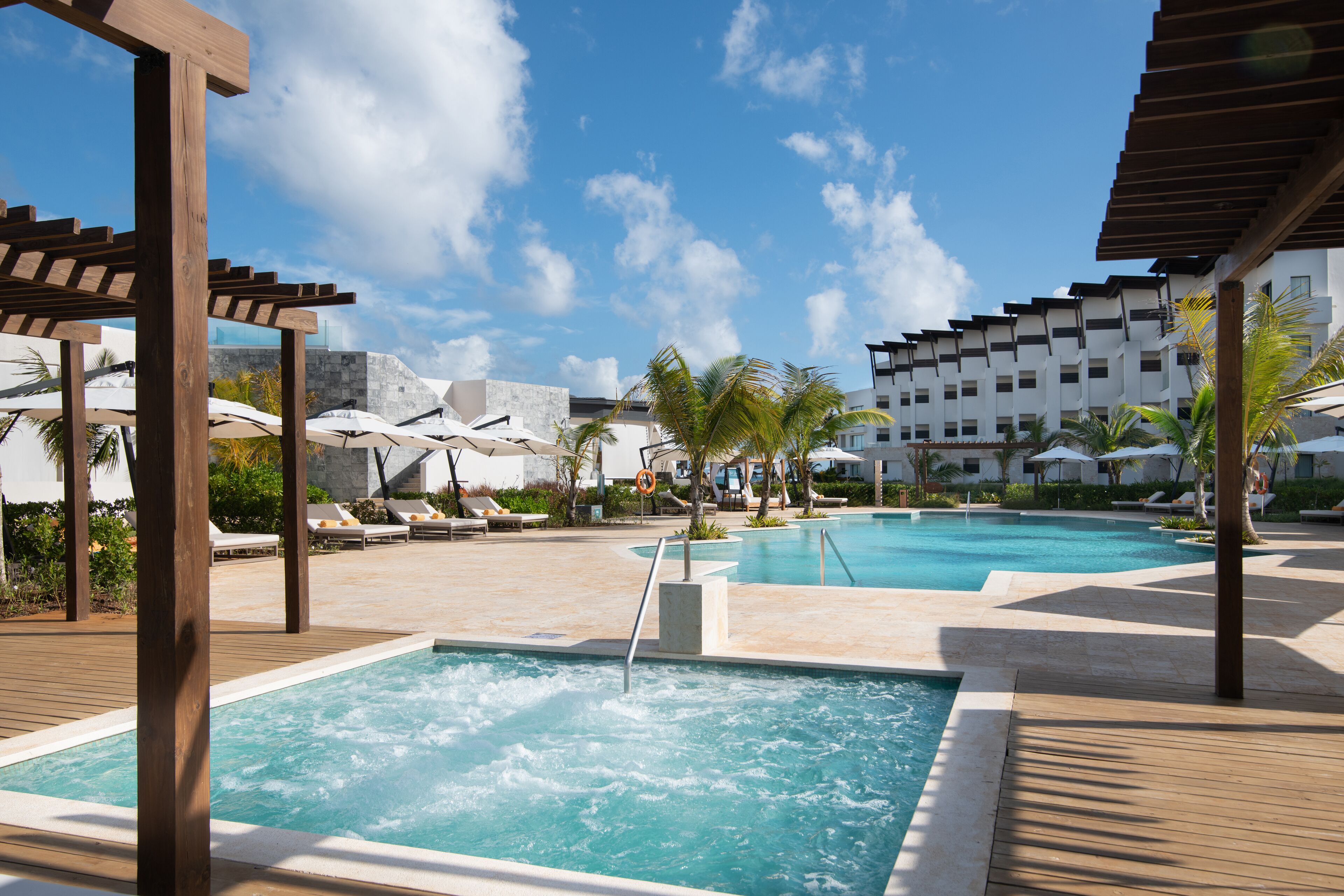 A pool area with two separate pools, lounge chairs, umbrellas, and a wooden pergola structure.