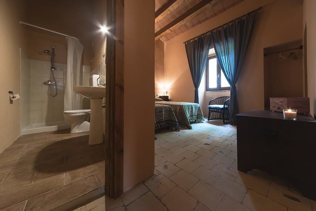 A bathroom with a shower, toilet, and sink. There is a bed with a blanket and a bedside table with a candle. The room has a wooden floor and a window with curtains.