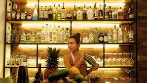 Uma área de bar com prateleiras cheias de garrafas de bebidas, um bartender preparando uma bebida e uma exposição de frutas no balcão.