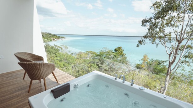 Uma banheira de hidromassagem numa varanda com vista para uma praia, água azul cristalina e vegetação.