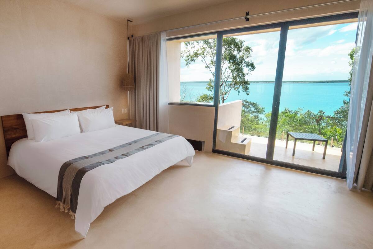 A bedroom with a large bed, a wooden nightstand, a desk, and a view of the sea through a sliding glass door.