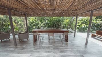 An outdoor covered patio with a wooden table and chairs, surrounded by wicker furniture and a view of lush greenery.