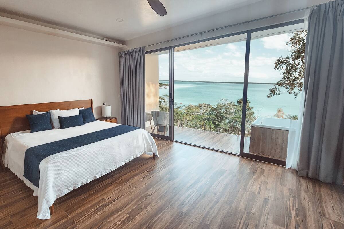 A bedroom with a large bed, wooden flooring, a nightstand, and a view of the ocean through a sliding glass door.