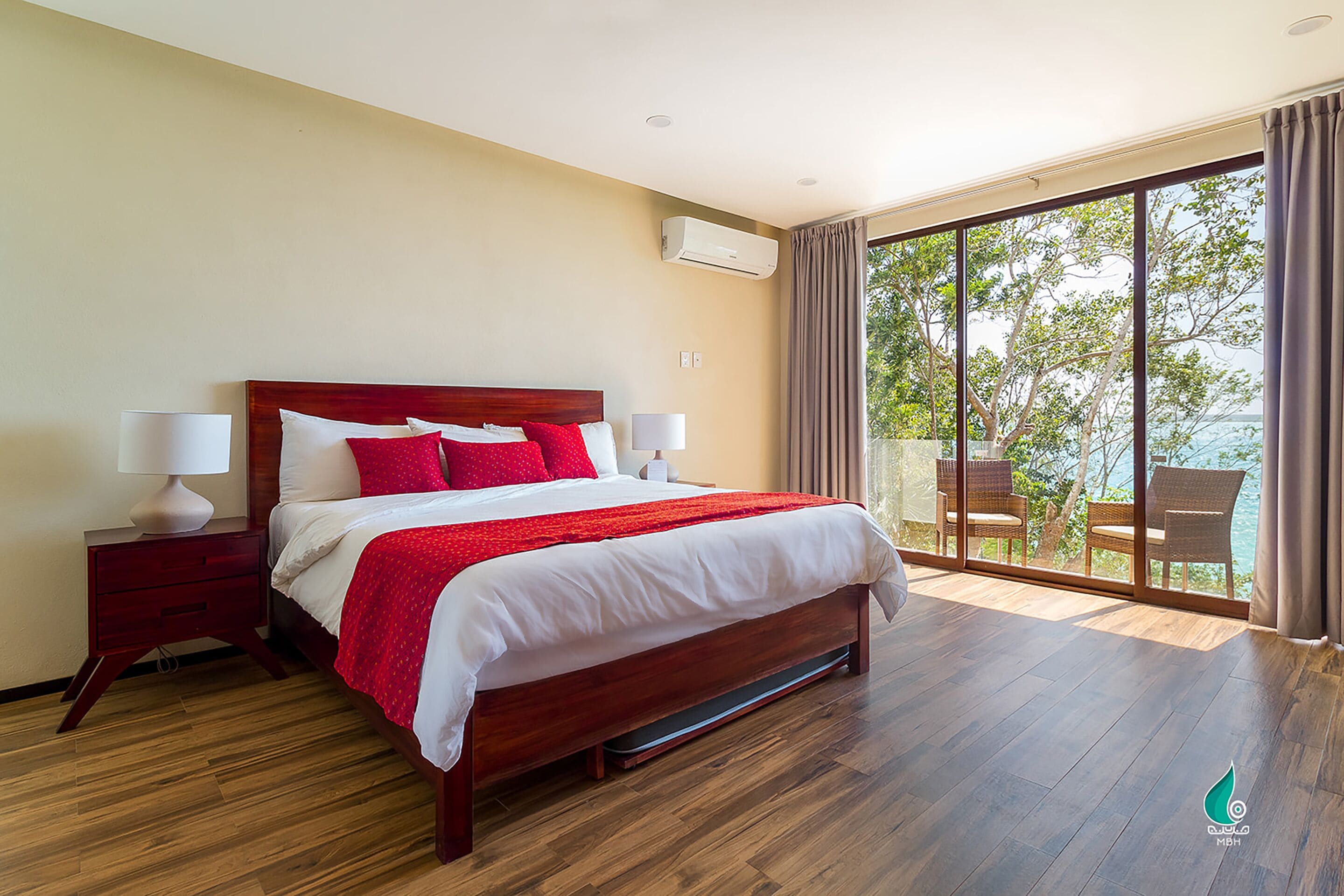 A spacious bedroom with a large bed, wooden floor, and a balcony with wicker chairs.