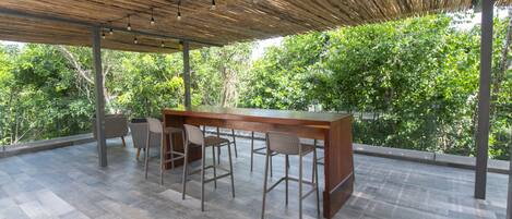 Une terrasse extérieure dotée d’un bar en bois et d’un espace salon, le tout entouré d’une végétation luxuriante.