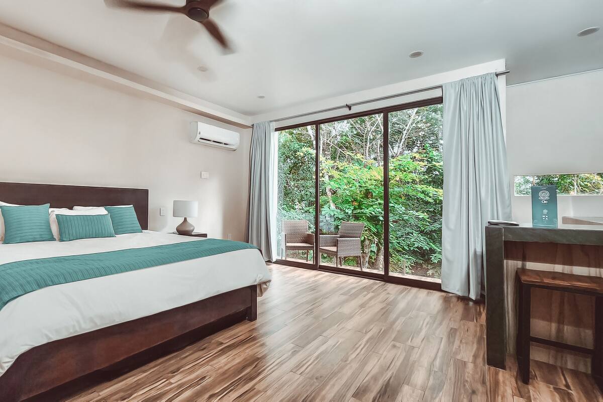 A modern hotel room with a large bed, a ceiling fan, a desk, and a view of the outdoors.
