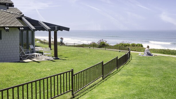 A coastal view with a grassy area, a building with a patio, and a fence. - WCH at Wave Crest Resort (Del Mar)