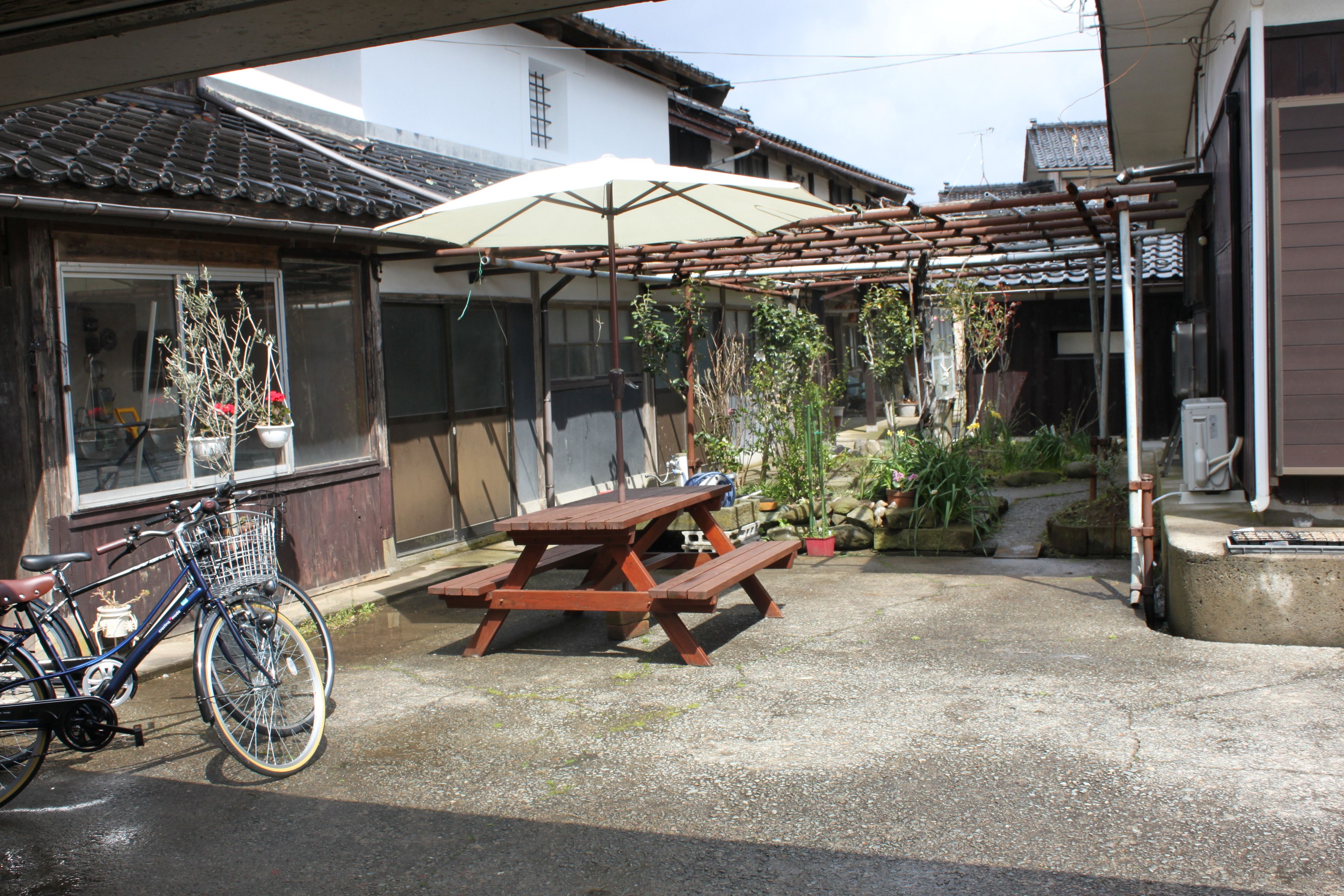 A wooden picnic table under a pergola with a white umbrella, a bicycle, and potted plants in a residential setting.