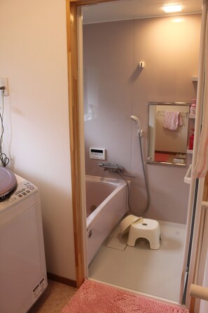 A small bathroom with a bathtub, a toilet, and a shower. There is a pink rug on the floor and a small stool next to the toilet.