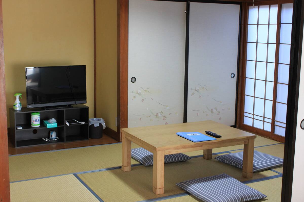 A traditional Japanese room with tatami flooring, a low wooden table, cushions, a television on a shelf, and sliding shoji doors.