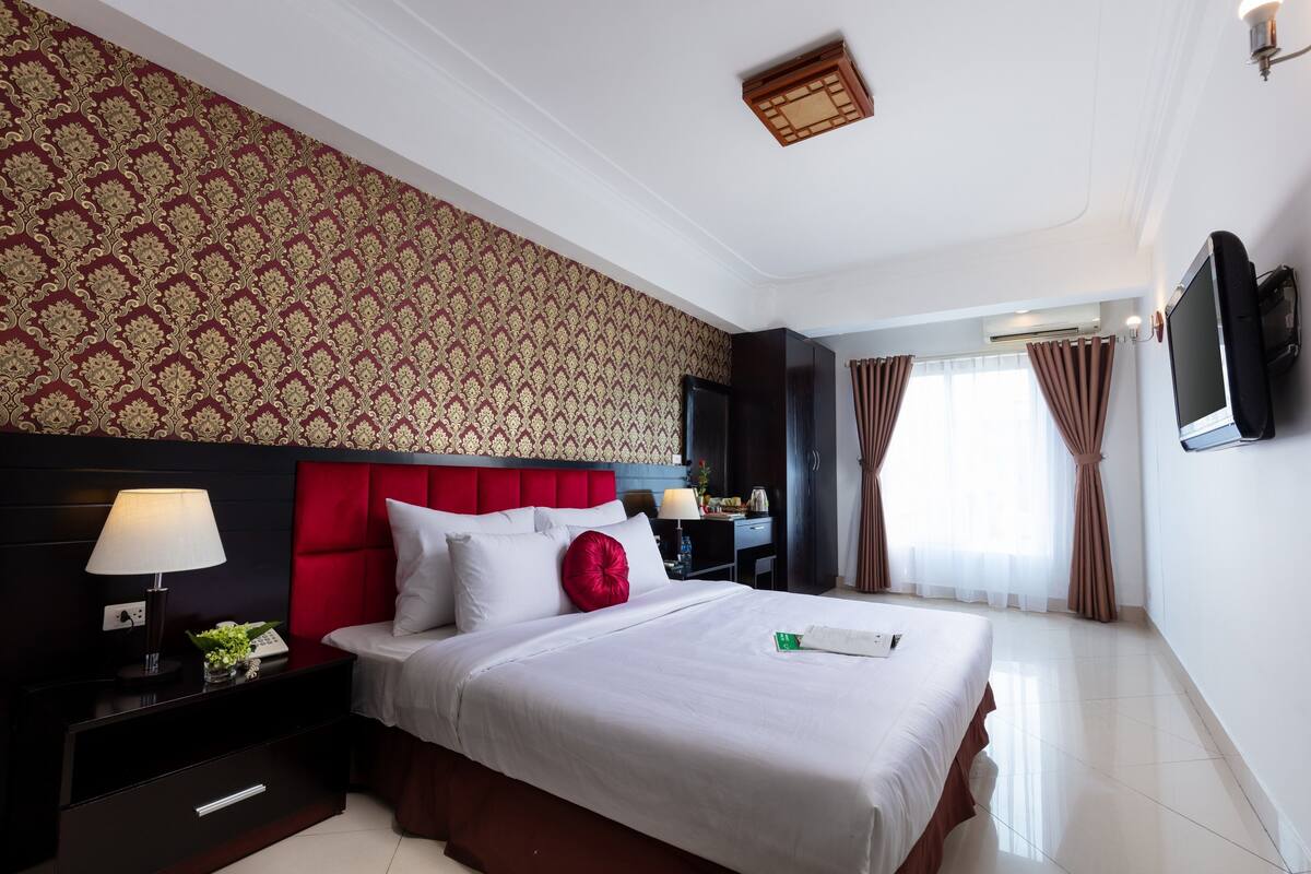 Une chambre d’hôtel avec un grand lit, une tête de lit rouge, une table de chevet avec une lampe, un bureau avec un ordinateur et un téléviseur fixé au mur.