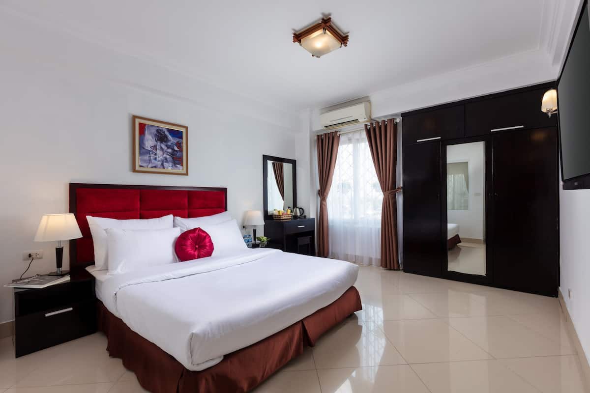 Une chambre d’hôtel avec un grand lit, une tête de lit rouge, un téléviseur à écran plat, une table de chevet avec une lampe et une fenêtre avec des rideaux.