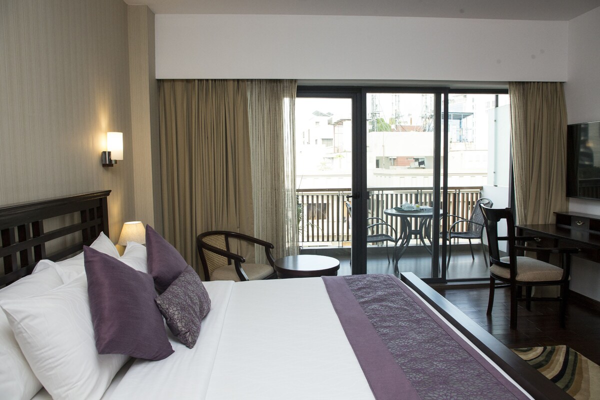 Habitación de hotel con cama, escritorio, silla, mesa y balcón con vista a edificios.