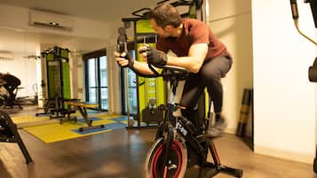 Un homme fait de l’exercice sur un vélo d’appartement dans une salle de sport, où l’on trouve également d’autres appareils d’entraînement et des tapis au sol.