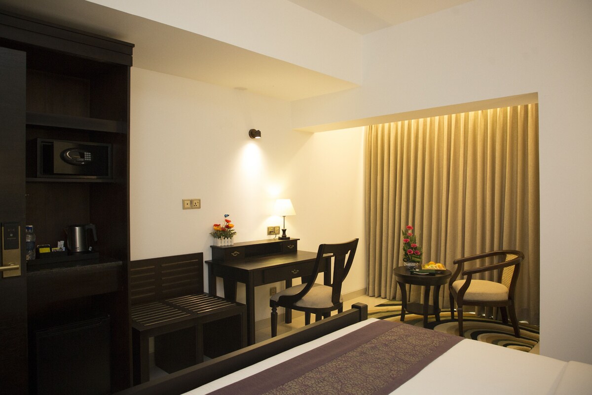 Habitación de hotel con cama, escritorio, dos sillas, lámpara, microondas y cortina.