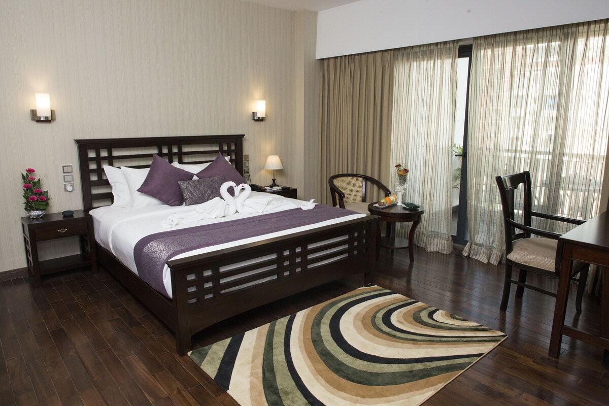 A hotel room with a large bed, a desk, and a chair. There is a window with curtains, a nightstand with a lamp, and a vase with flowers.
