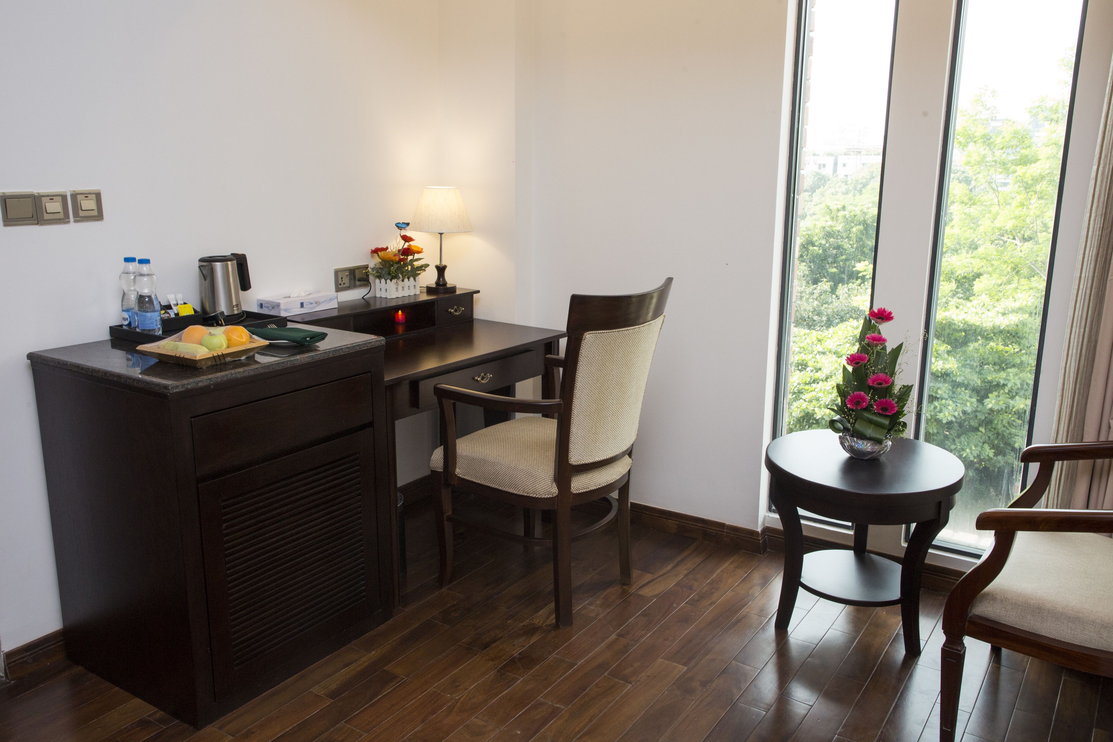 A small, dark wood-framed desk with a chair, a small round table with a vase of flowers, and a window with a view of trees.