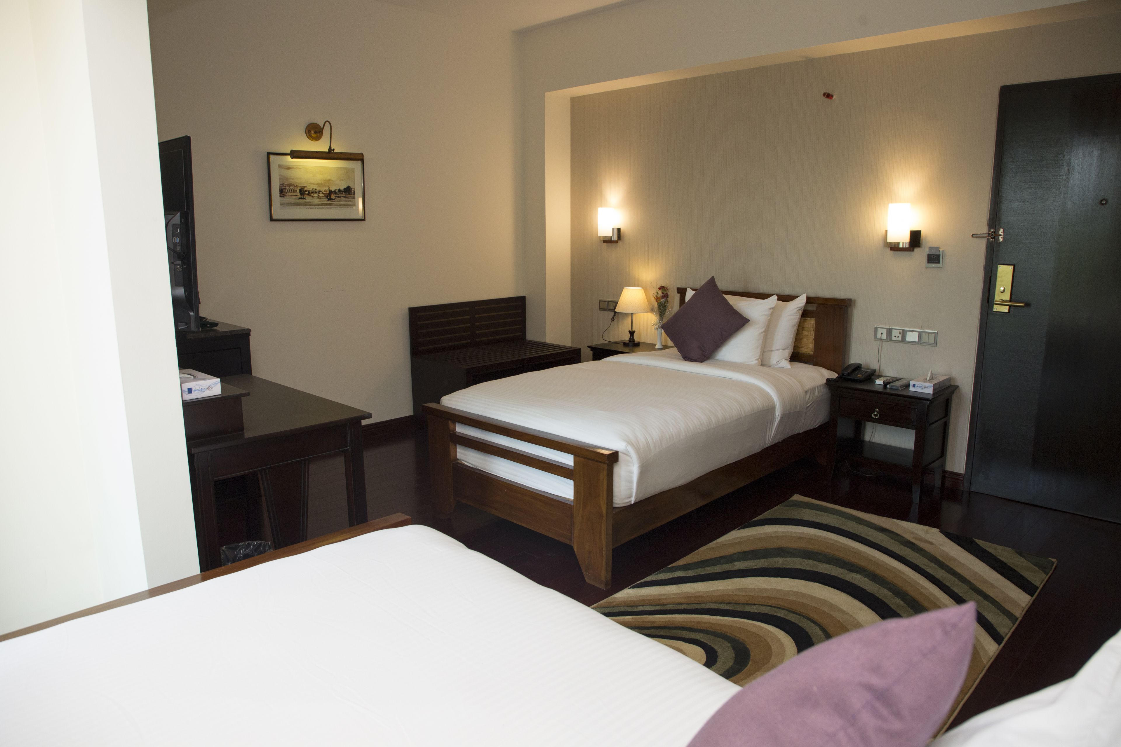 Quarto de hotel com uma cama grande, mesas de cabeceira, uma escrivaninha, uma cadeira, uma TV e um quadro na parede.