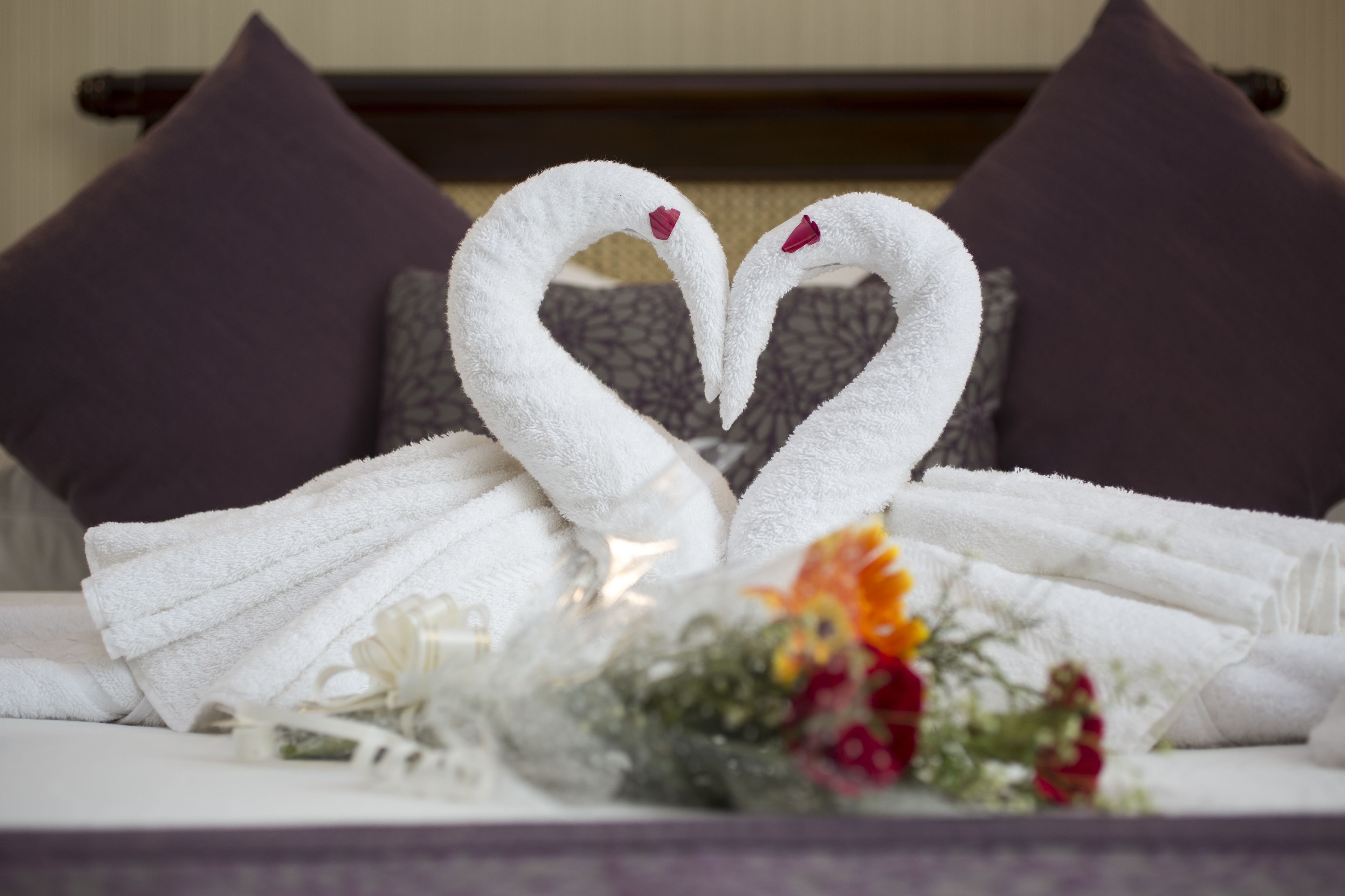 Uma cama com duas toalhas brancas dispostas em formato de cisnes com corações, um buquê de flores e um travesseiro com padrão geométrico.