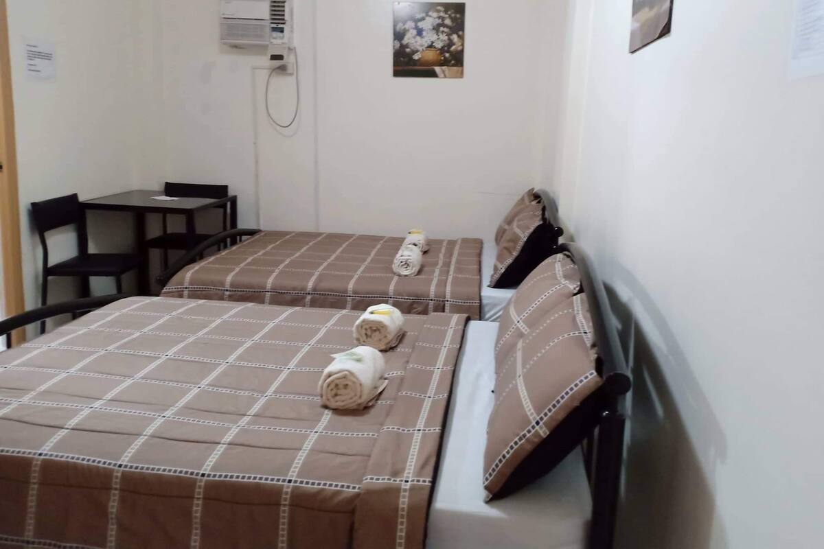 A hotel room with two beds, each with a brown checkered bedspread and pillows, a small table, and a chair.