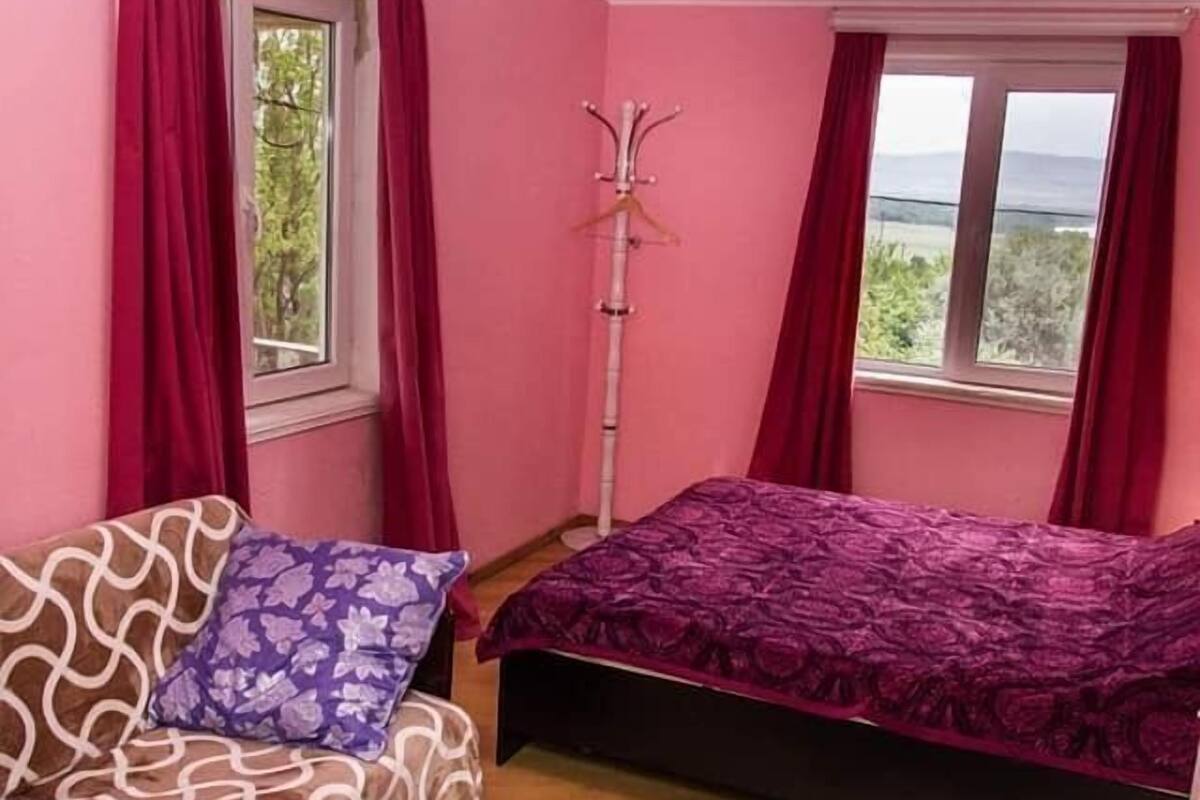 A room with pink walls, a bed with a purple bedspread, a sofa with a patterned cushion, and a wooden floor.