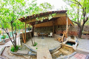 A rustic outdoor seating area with a wooden pergola, stone flooring, and a small fountain. - Amiran`s Lake (Tbilisi)