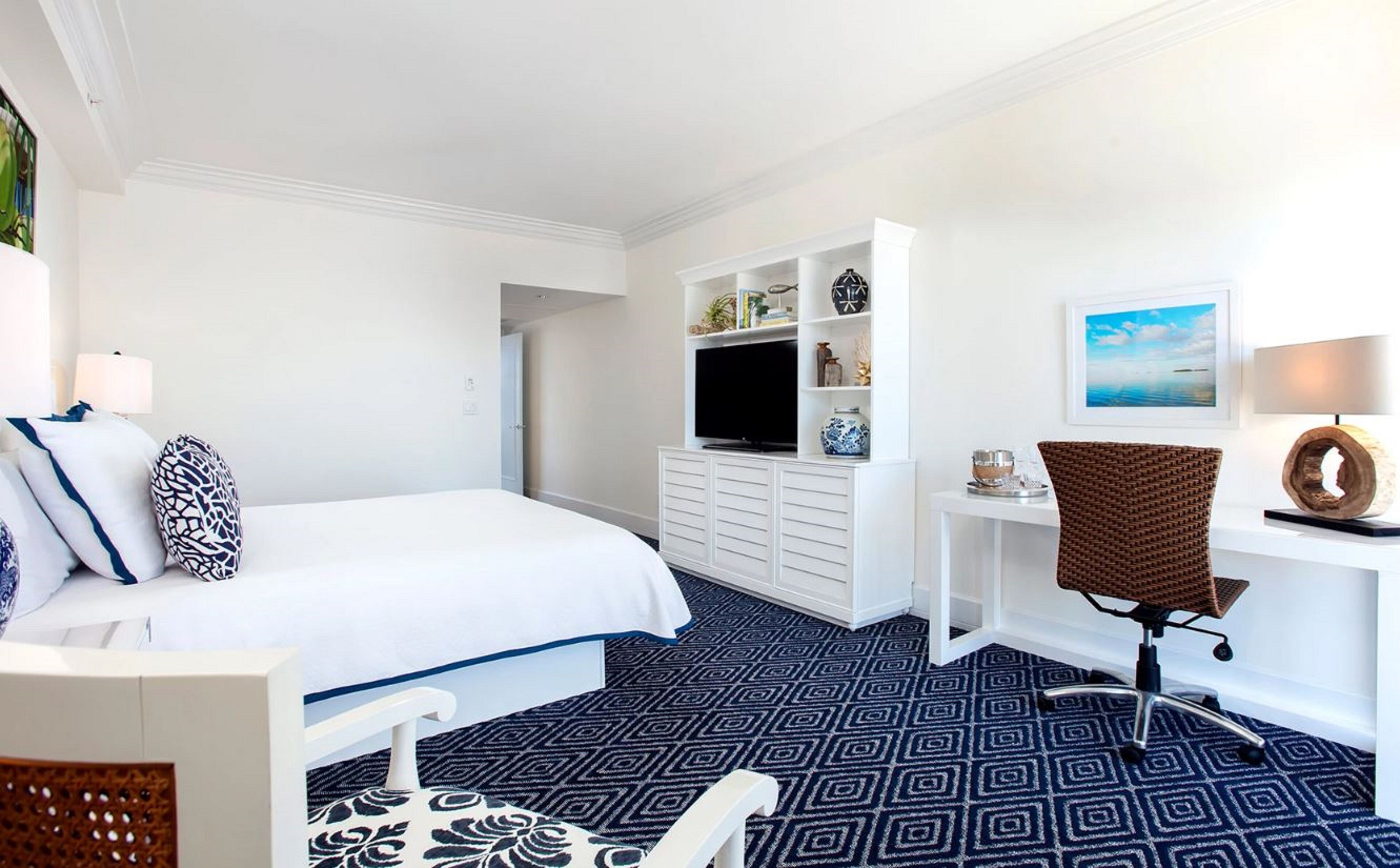A hotel room with a bed, a desk, a chair, a TV, and a painting on the wall.