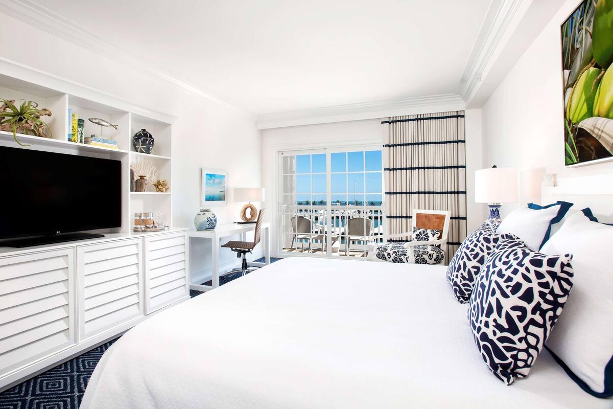 Habitación de hotel con una cama grande, televisor de pantalla plana, estantería, zona de comedor con sillas y vista al océano.