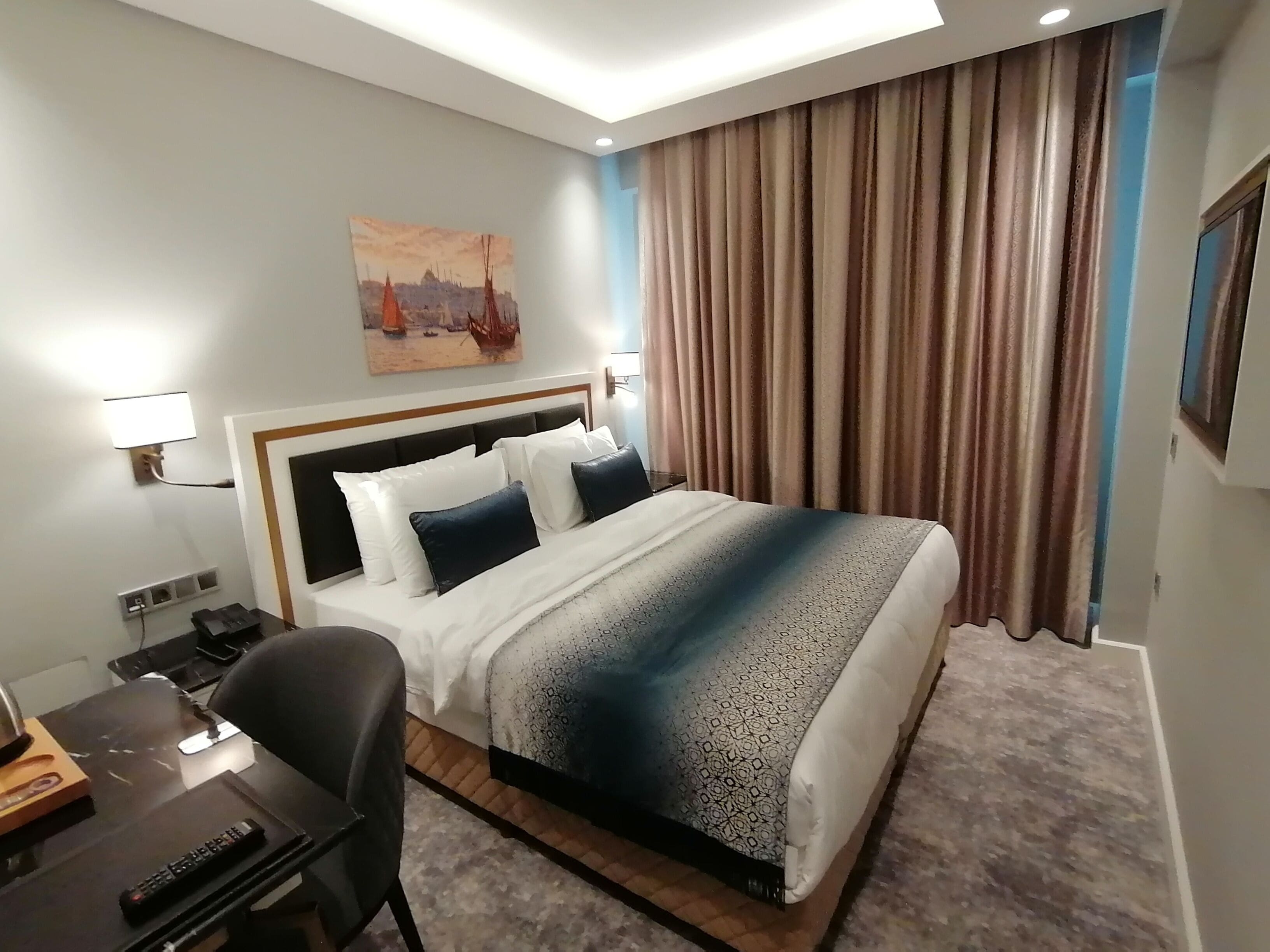 Habitación de hotel con cama, escritorio, silla, un cuadro en la pared y ventana con cortinas.