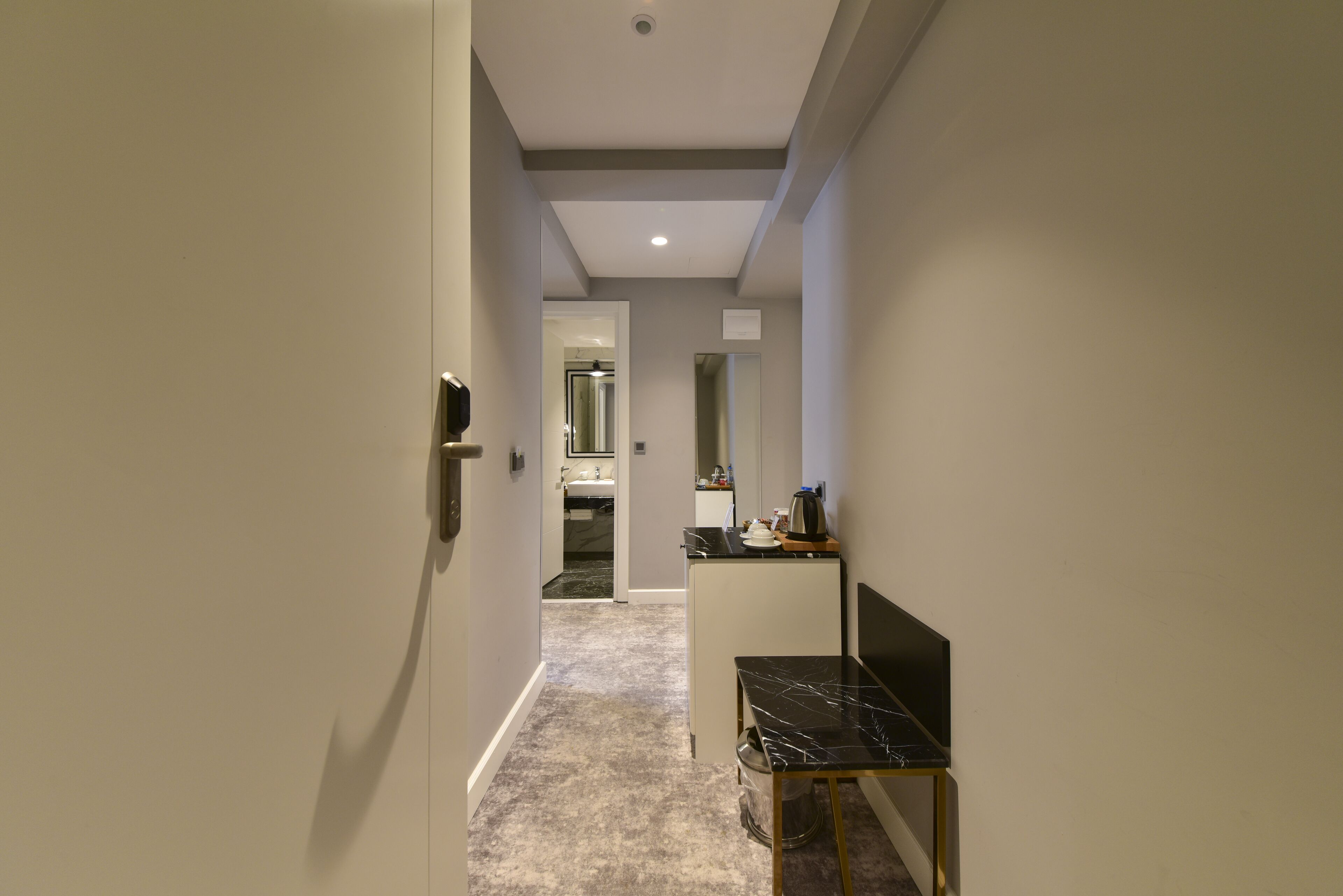 A narrow hallway with a door on the left, a small table with a black marble top and a trash can on the right, and a kitchen area in the background.